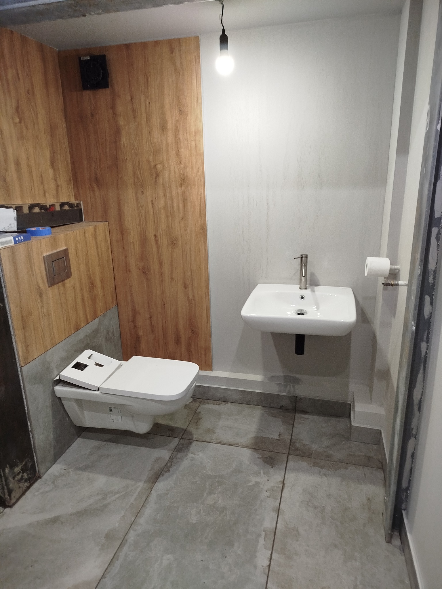 Nowoczesna łazienka z podwieszaną toaletą i umywalką. Ściany wykończone panelami drewnopodobnymi i jasnymi płytkami. Minimalistyczny design z surowym betonem na podłodze.