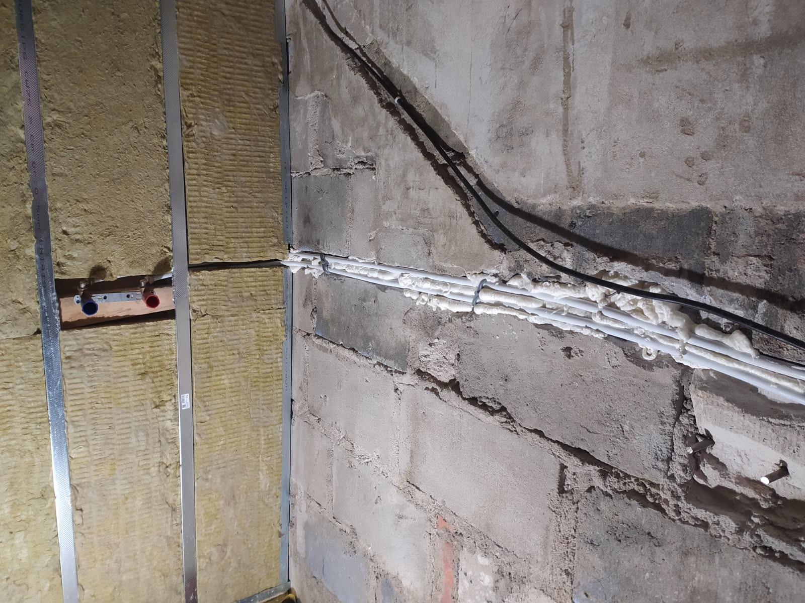 Ocieplenie ściany wełną mineralną na ruszcie z profili stalowych, widoczne rury instalacyjne zabezpieczone pianką montażową, surowa ściana z betonu komórkowego.