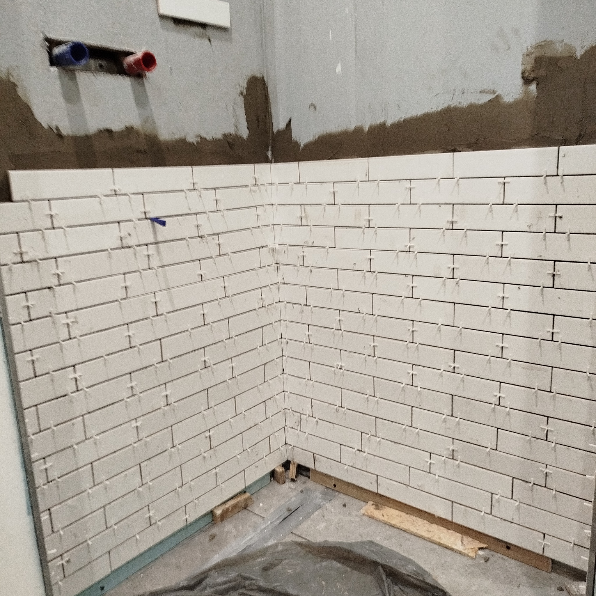 Ściana wyłożona białymi, prostokątnymi płytkami, z widocznymi krzyżykami dystansowymi. Fragment ściany z instalacją wodną w tle. Prace glazurnicze w trakcie realizacji.