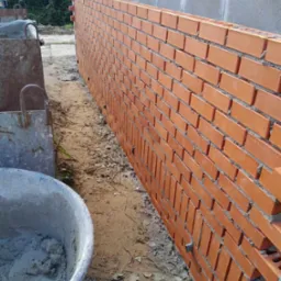 Wznoszona ściana z czerwonej cegły na fundamencie z szarego bloczka betonowego, widoczna zaprawa murarska i wiadro z cementem.