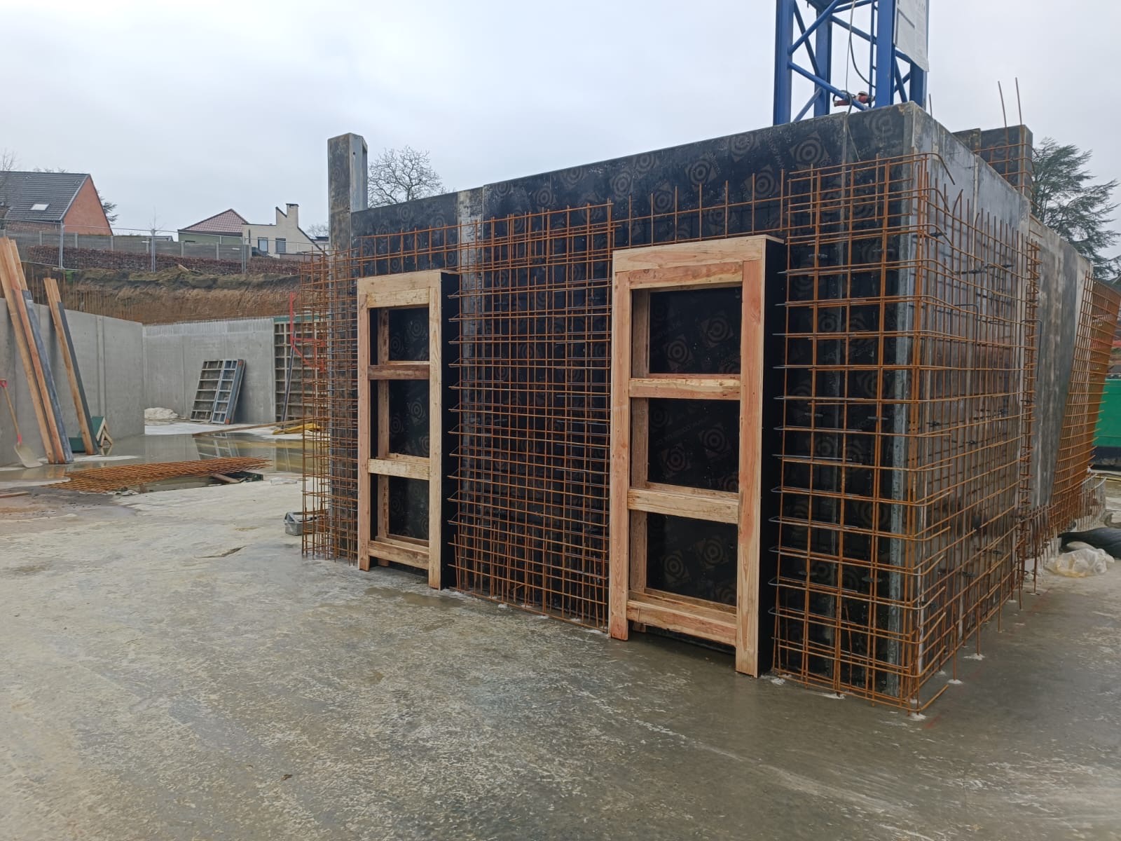 Zbrojenie fundamentów z drewnianymi ramami okiennymi na tle czarnej hydroizolacji w trakcie budowy domu, widoczny dźwig i teren budowy.