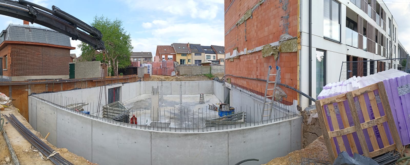 Wykop fundamentowy z betonowymi ścianami, widoczne zbrojenia, palety z materiałami izolacyjnymi oraz fragment budynku w trakcie budowy z ociepleniem na ścianach.