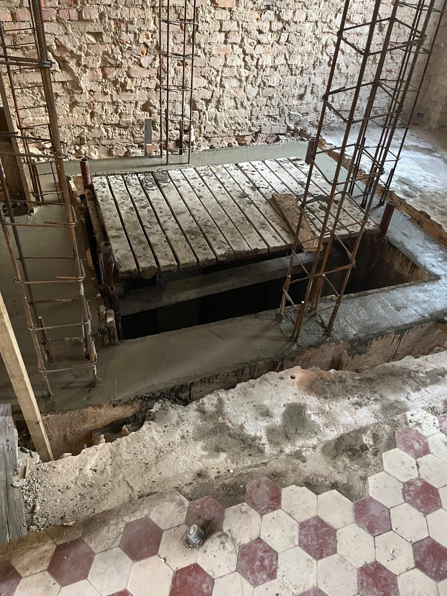 Wykop zbrojony stalowymi prętami i zalany betonem, widoczne drewniane szalunki, fragment ceglanej ściany i posadzki z heksagonalnych płytek w odcieniach czerwieni i bieli.