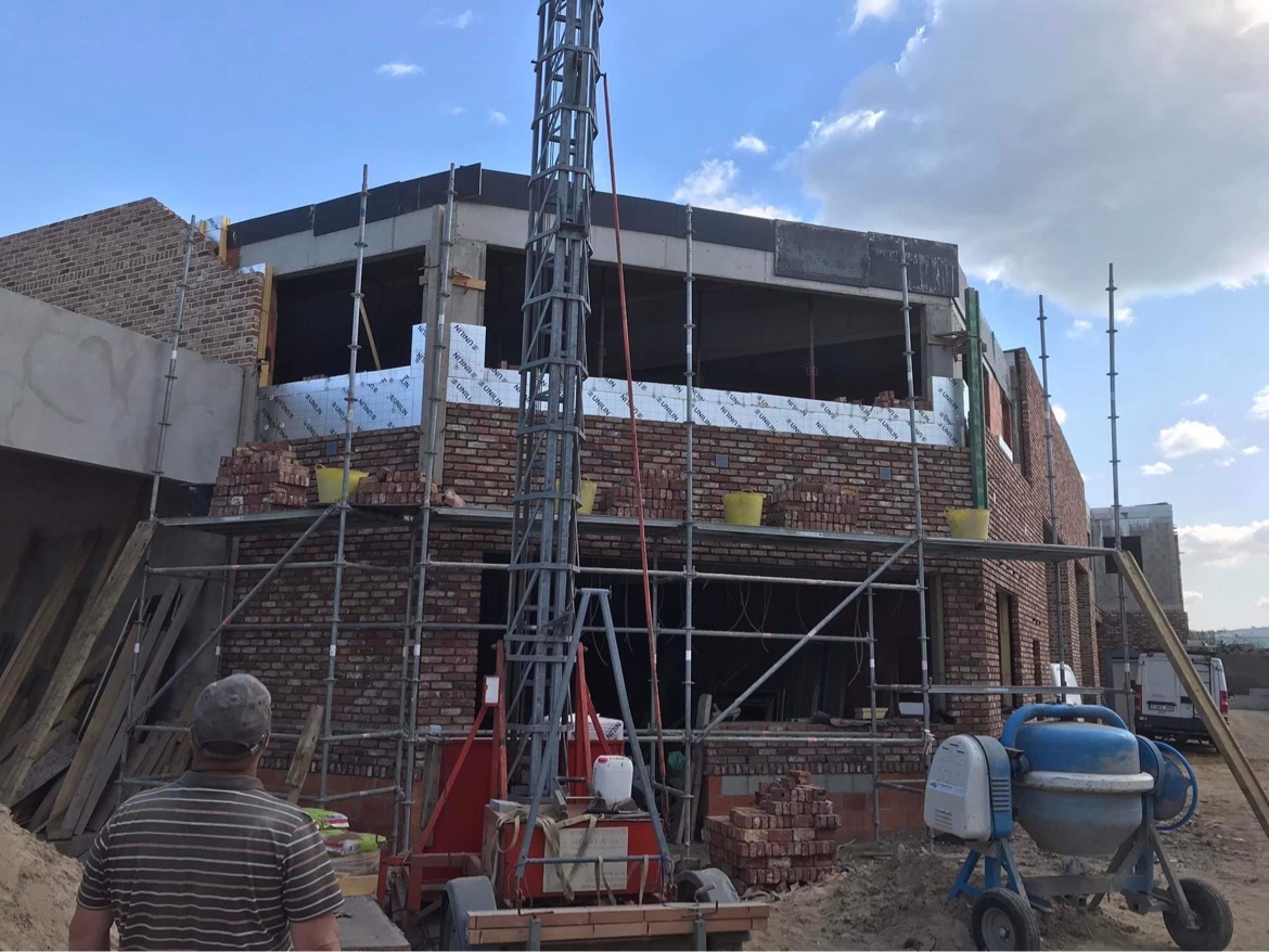Budowa domu z czerwonej cegły w trakcie realizacji, widoczne rusztowania, materiały izolacyjne, betoniarka i robotnik z tyłu.