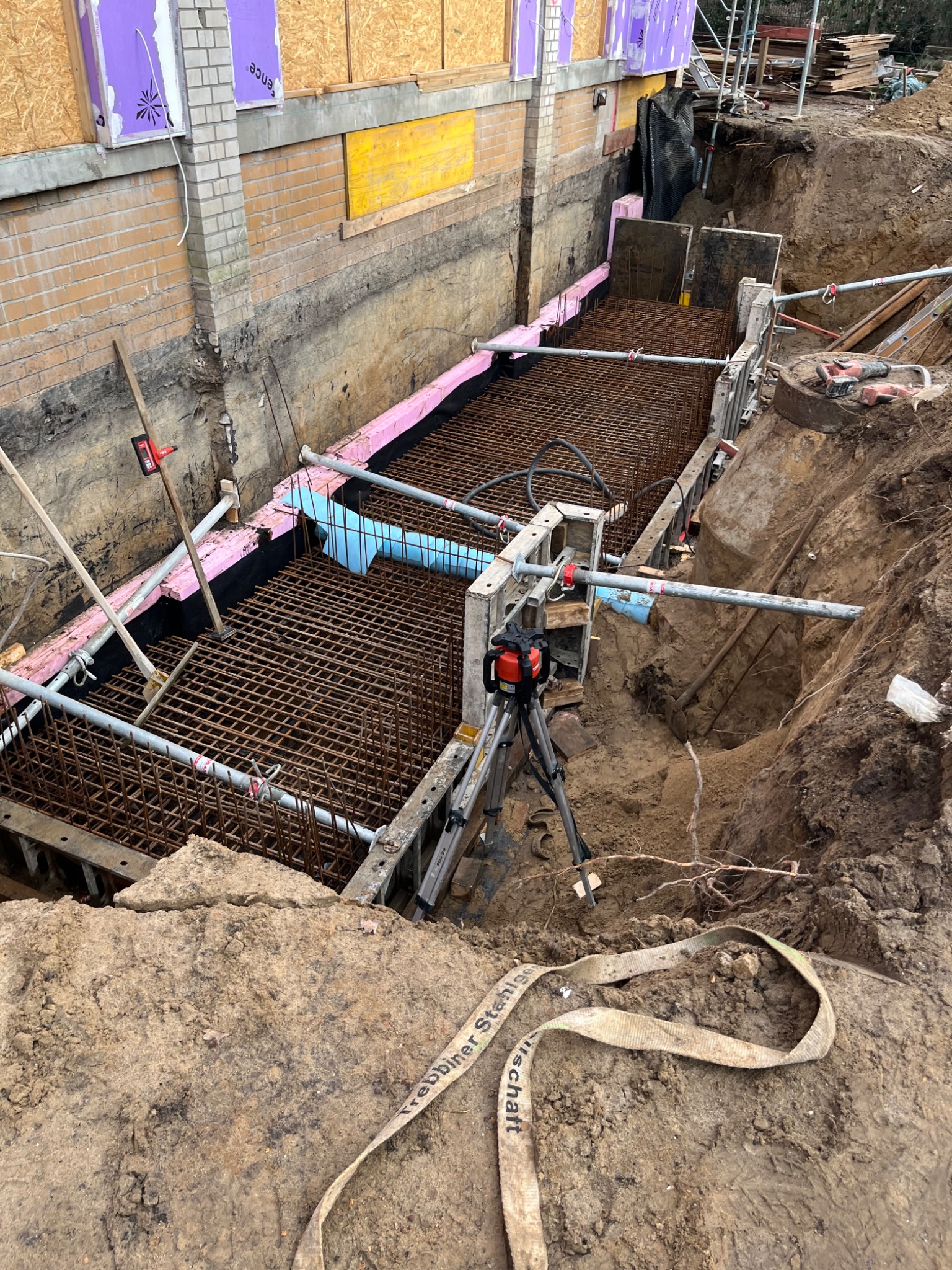 Zbrojenie fundamentów w wykopie, wzmocnione szalunkami, z widoczną izolacją pionową i poziomą, przygotowane do zalania betonem, z laserowym niwelatorem na statywie.