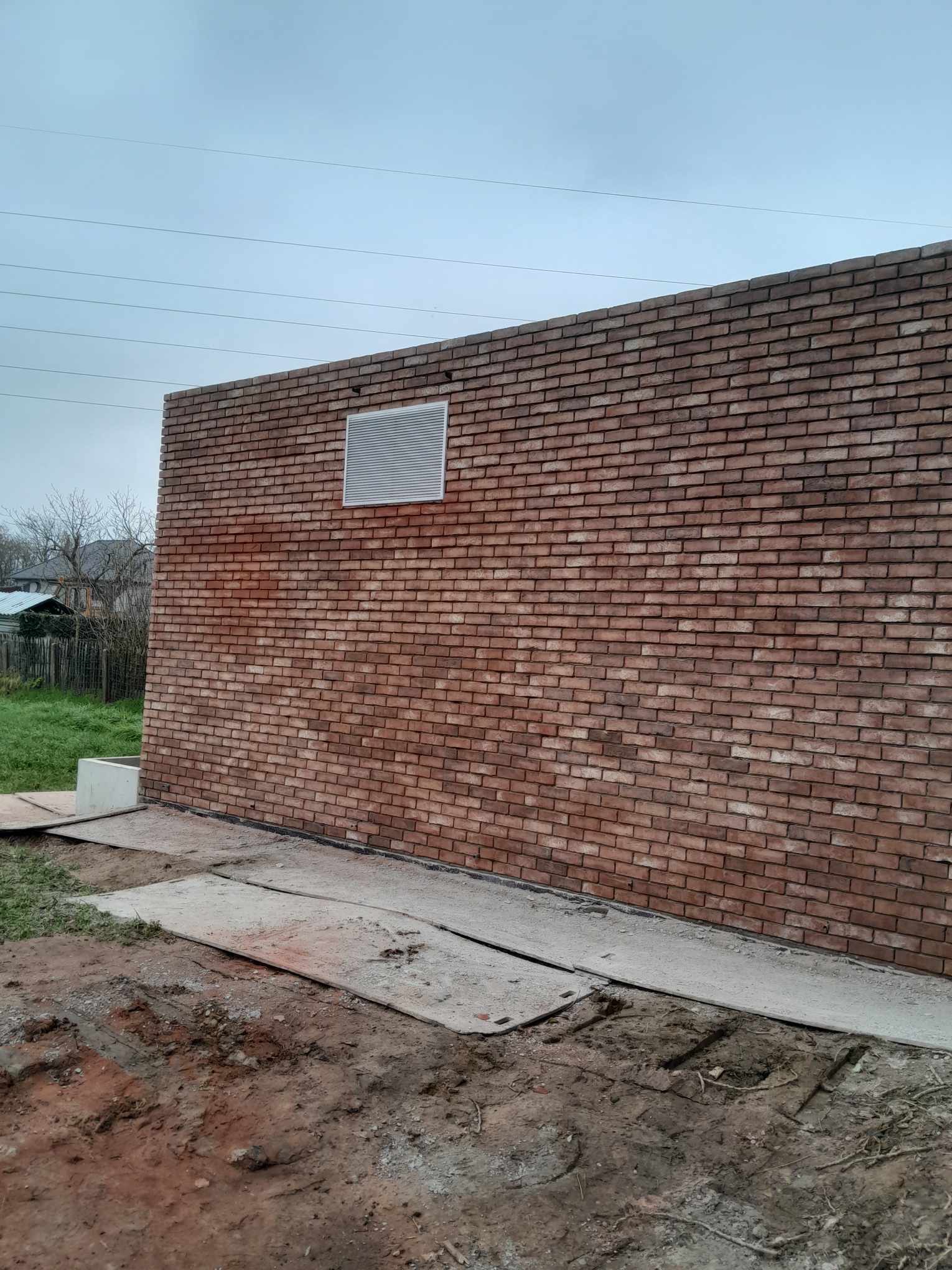 Ściana budynku z czerwonej cegły z kratką wentylacyjną, widoczne płyty chodnikowe i przygotowany teren pod trawnik.