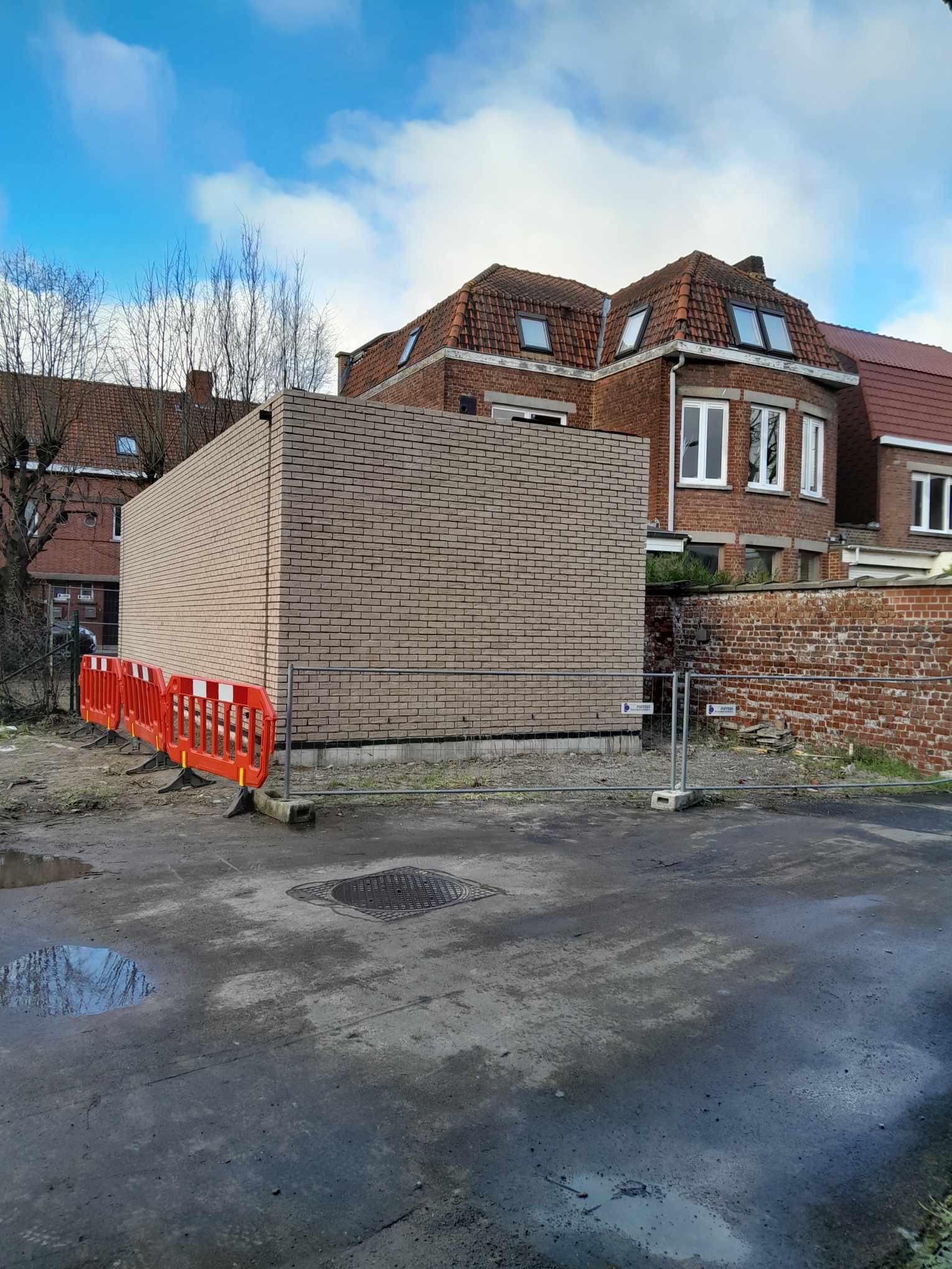 Wzniesiony fragment murowanej ściany z jasnej cegły, zabezpieczony pomarańczowymi barierkami budowlanymi, na tle zabudowań mieszkalnych z czerwonej cegły i pochmurnego nieba.