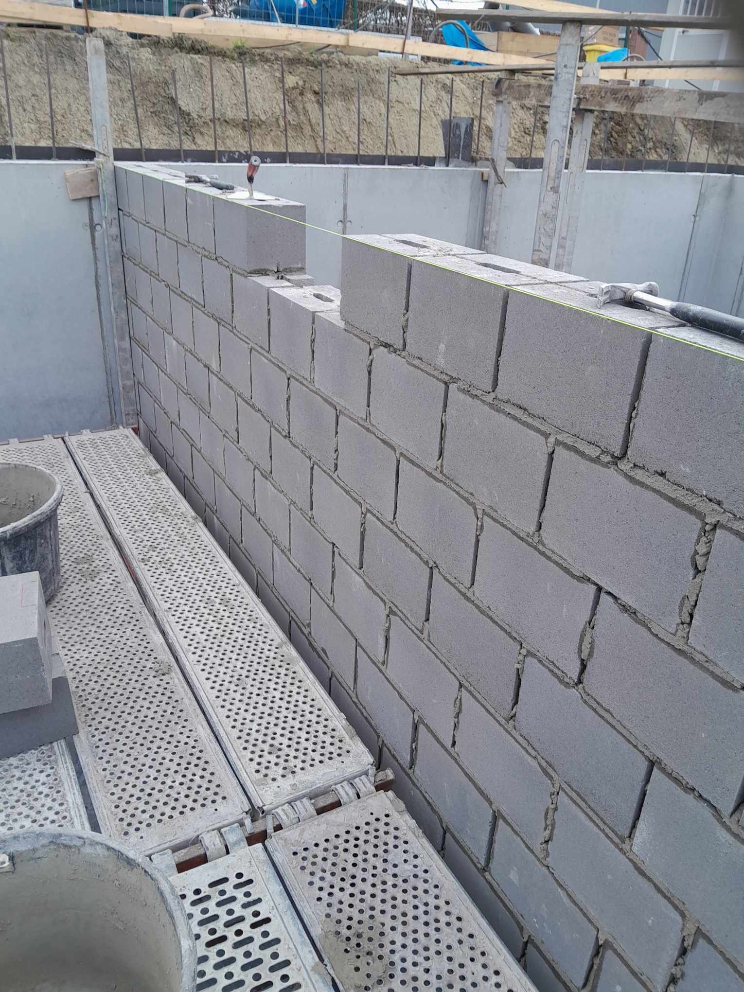 Wznoszenie ściany z bloczków betonowych na fundamencie, widoczne rusztowanie i narzędzia murarskie, sznurek murarski wyznaczający poziom.