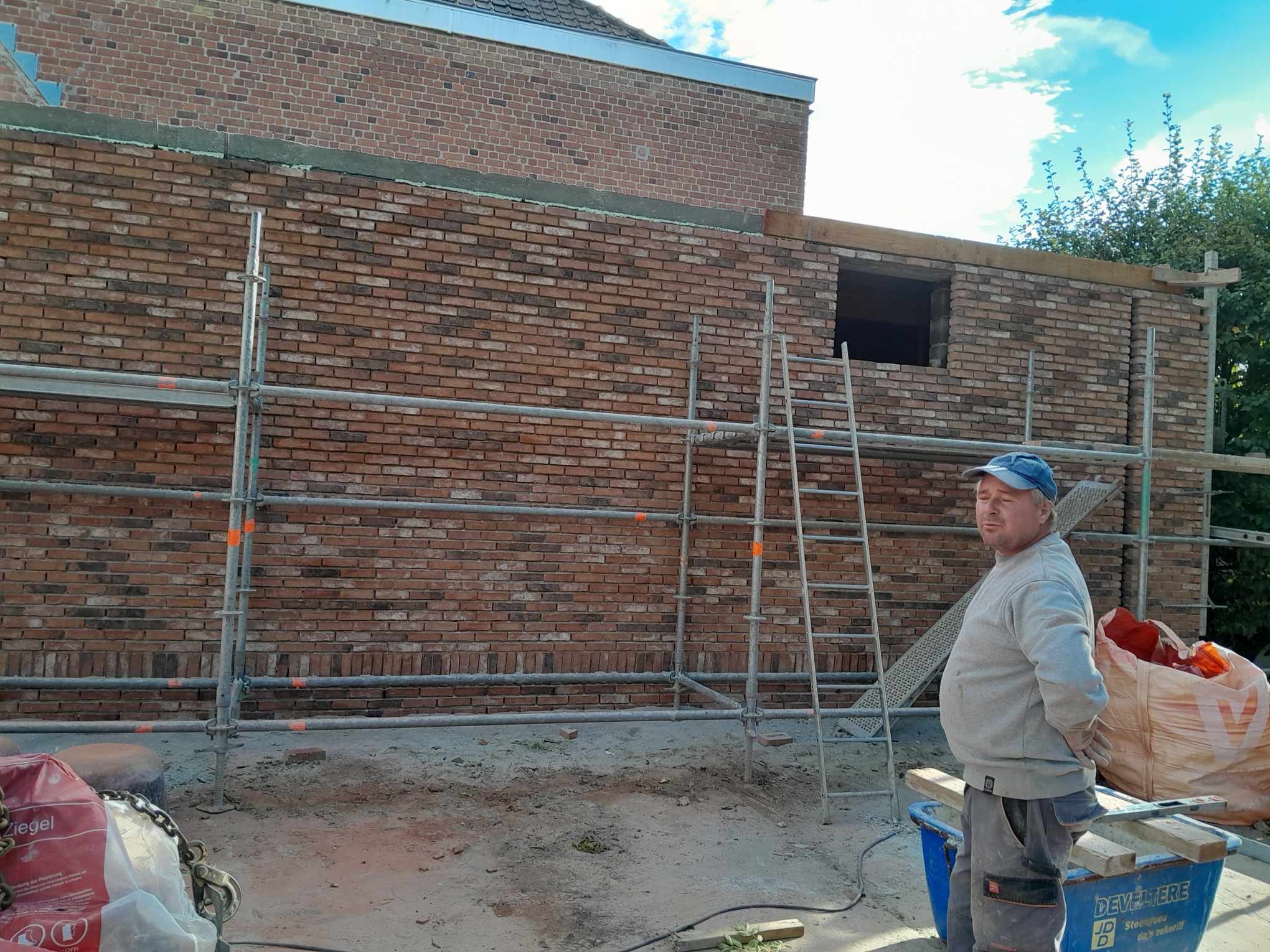 Murarz w niebieskiej czapce stoi na tle rusztowania i nowo wzniesionej ściany z cegły klinkierowej z widocznym otworem okiennym, w trakcie budowy domu.