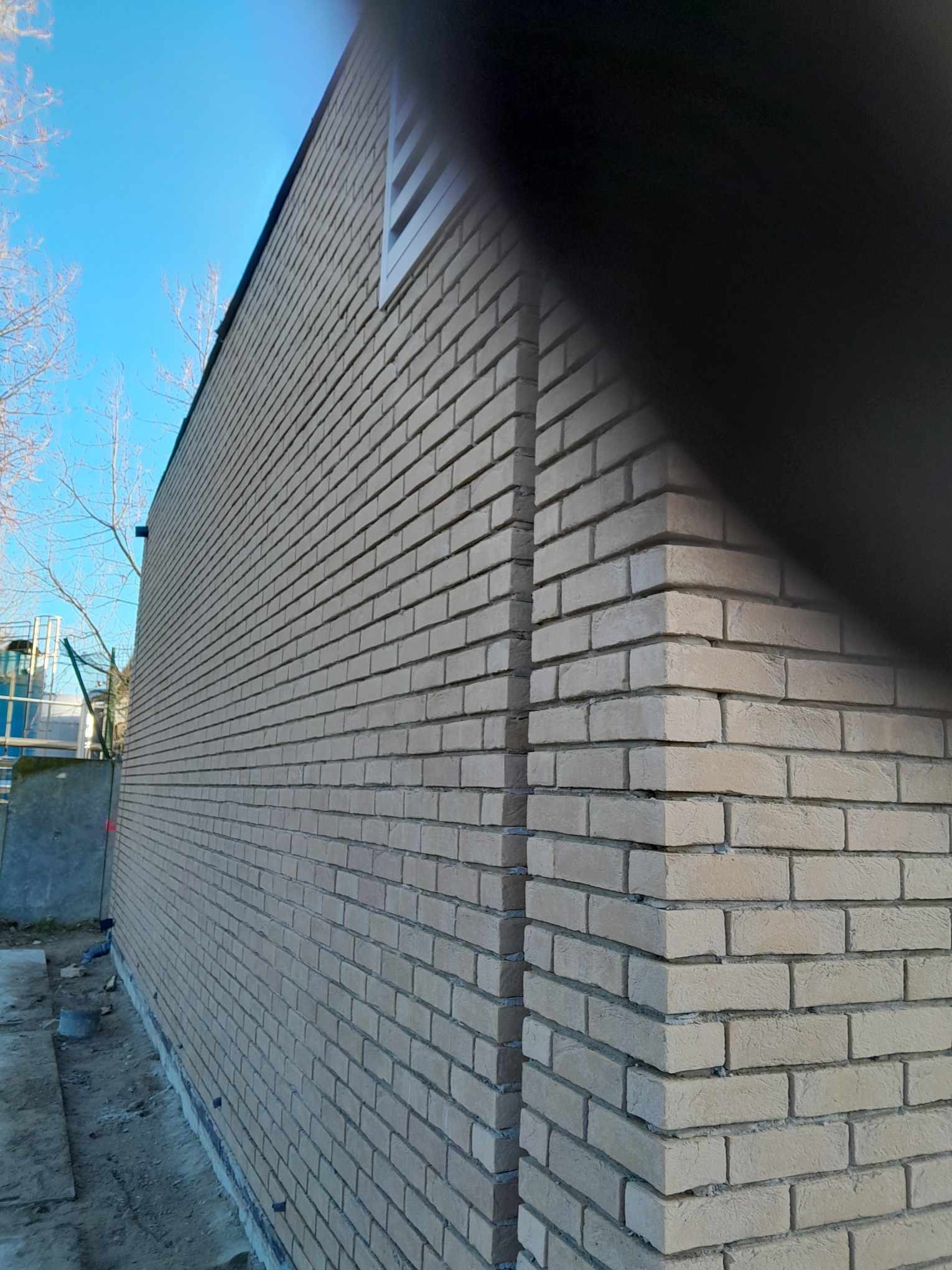 Świeżo wymurowana ściana zewnętrzna budynku z jasnej cegły, widoczny narożnik i fragment okna na tle błękitnego nieba.