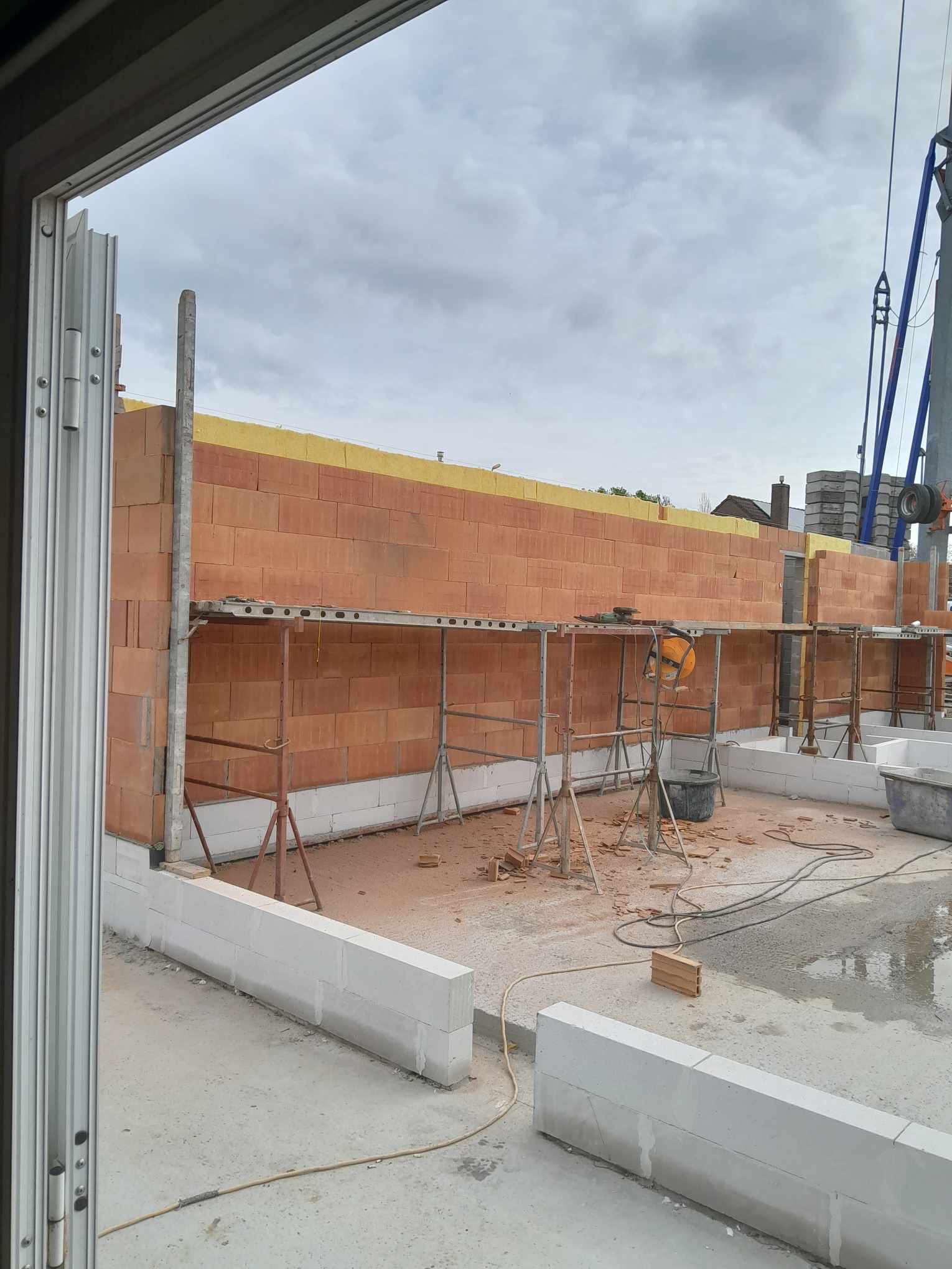 Wznoszenie ściany z czerwonej cegły na białym fundamencie, widoczne rusztowanie, żółta izolacja termiczna na górze ściany i dźwig budowlany w tle, widok z wnętrza budynku przez otwarte drzwi.