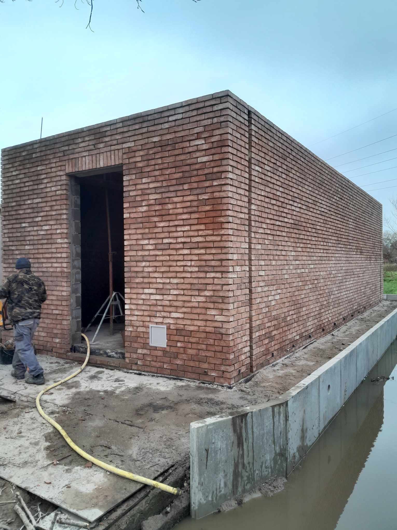 Surowa bryła budynku w trakcie budowy, wykonana z czerwonej cegły, widoczny pracownik z wężem i narzędziami, teren budowy nad kanałem wodnym.