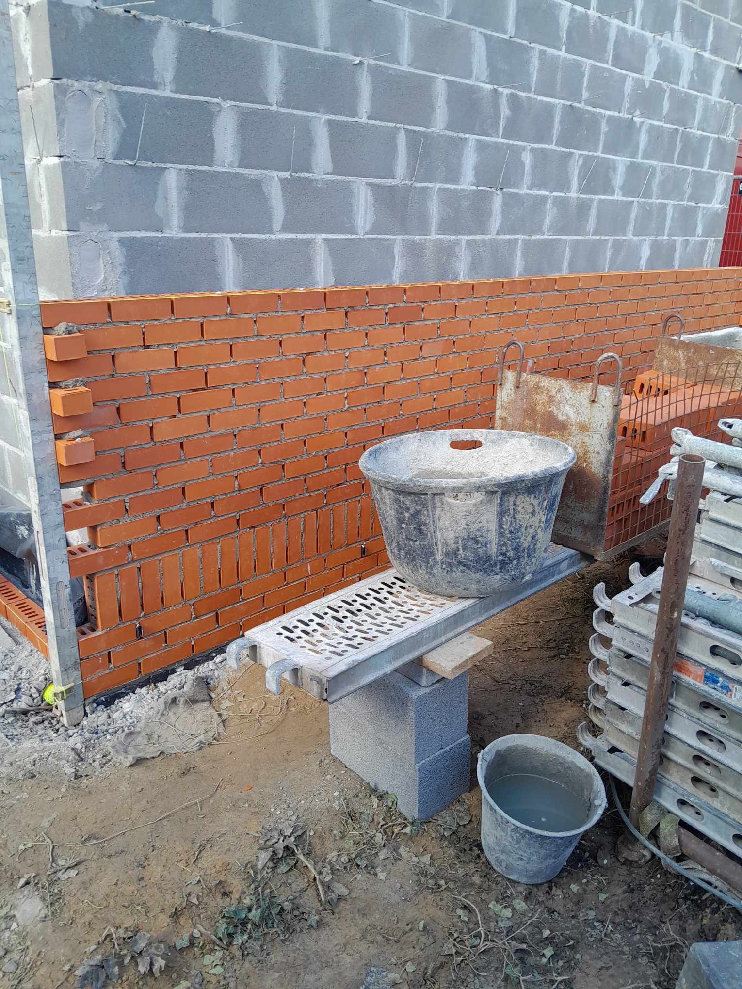 Murowanie ściany z cegły klinkierowej na tle ściany z bloczków betonowych, widoczne rusztowanie, wiadro z zaprawą i narzędzia murarskie.