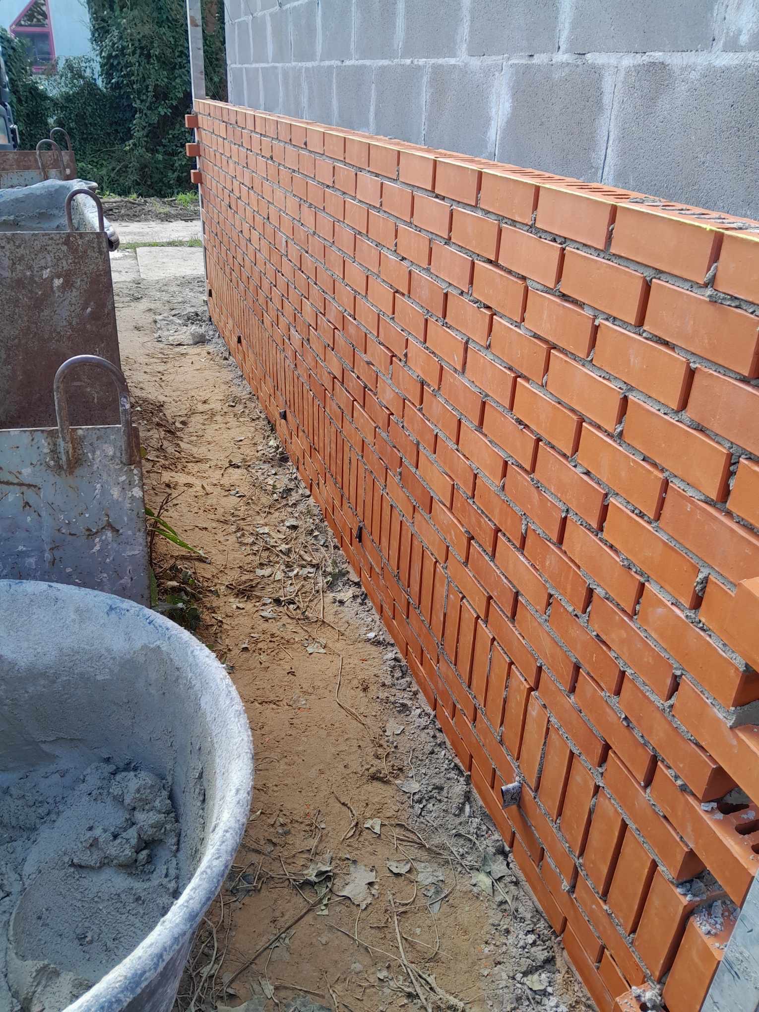 Wznoszona ściana z czerwonej cegły na fundamencie z szarego bloczka betonowego, widoczna zaprawa murarska i wiadro z cementem.