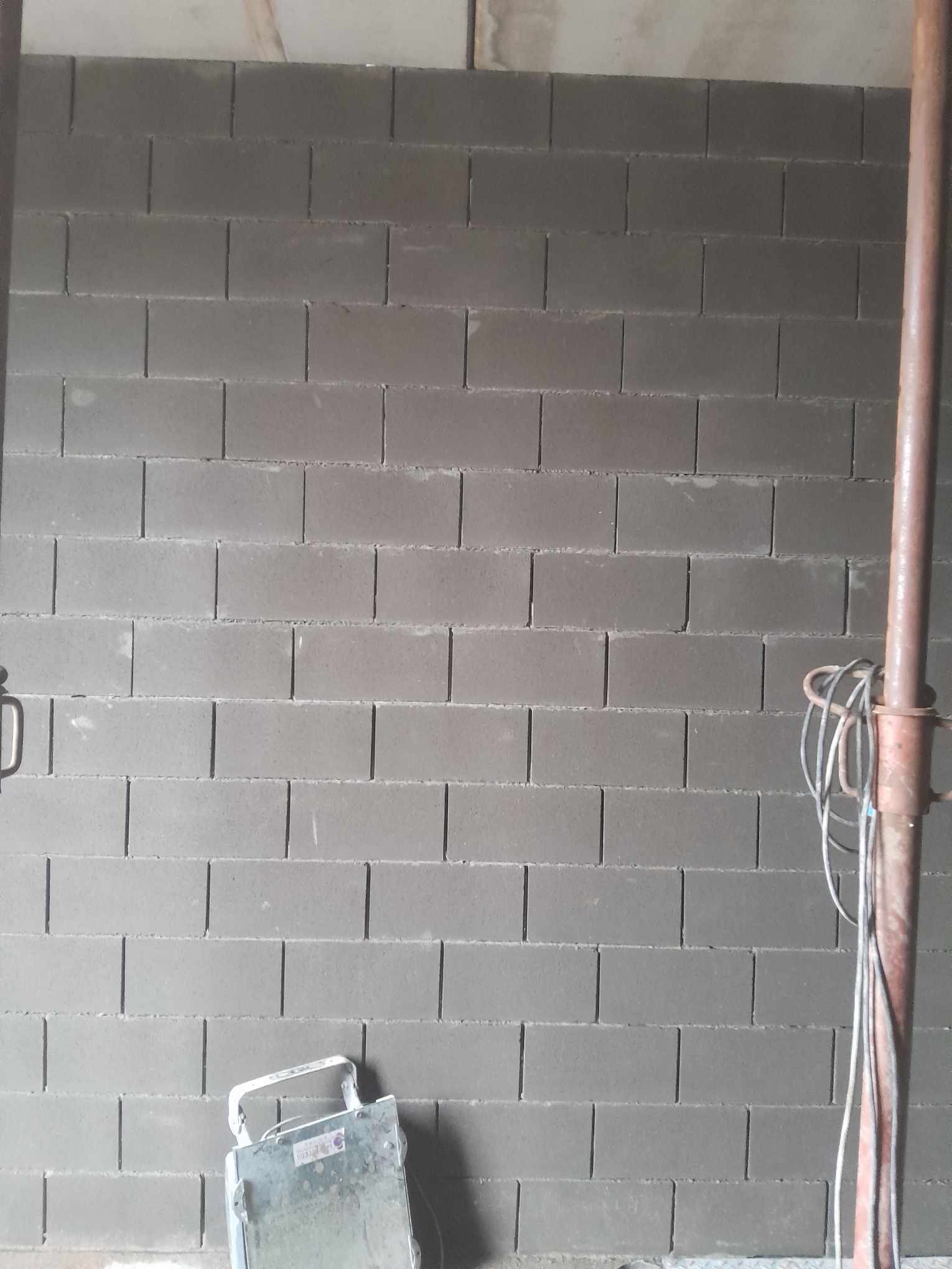 Świeżo wymurowana ściana z szarych bloczków betonowych; widoczny fragment metalowego rusztowania z kablami oraz metalowy podnośnik do płyt na podłodze.