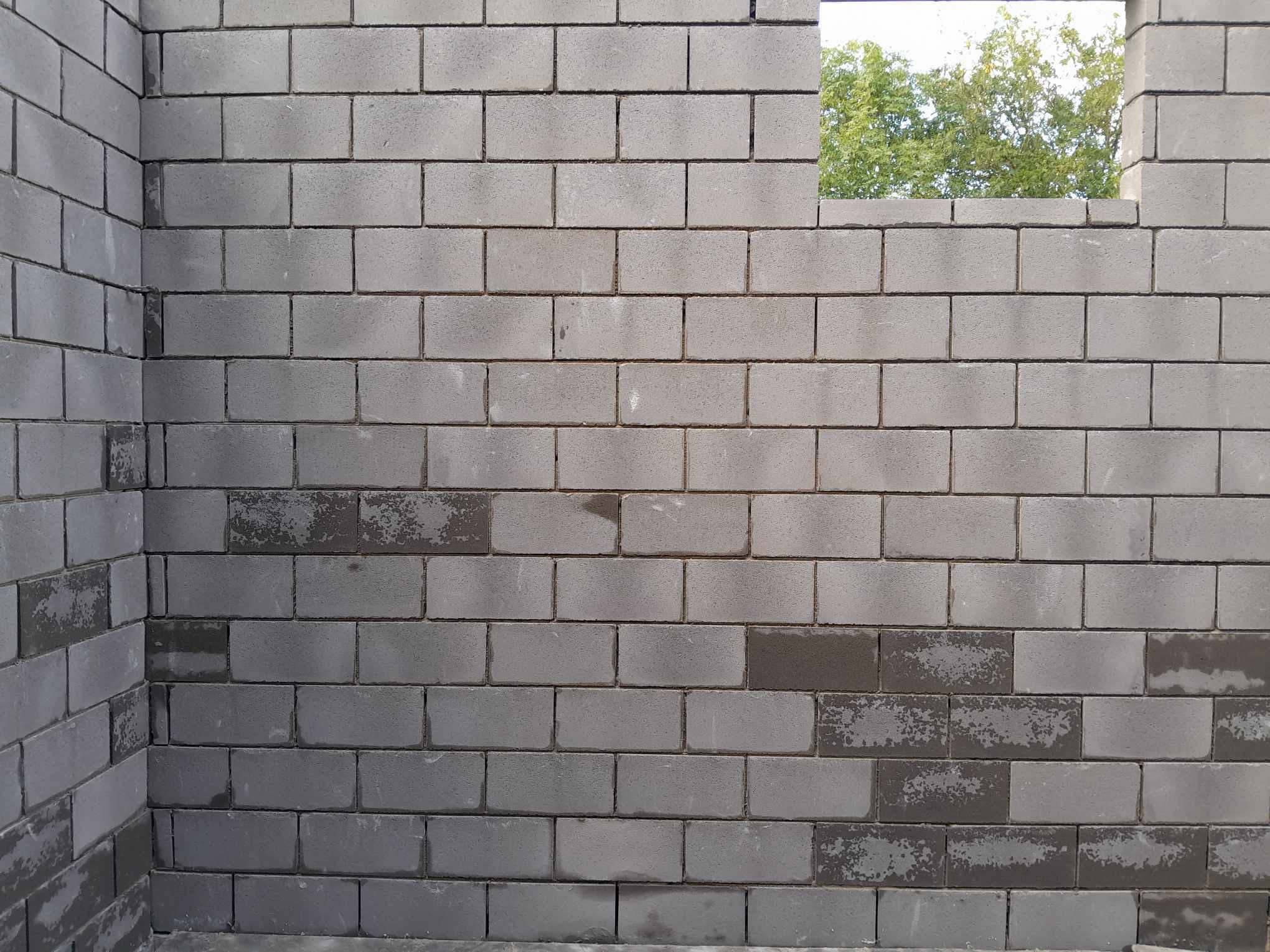 Wznoszenie ściany z szarych bloczków betonowych, widoczny narożnik oraz otwór okienny z zielenią w tle, ślady wilgoci na niektórych bloczkach.