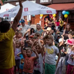 Animator w żółtej koszulce prowadzi grupę dzieci podczas zabawy na świeżym powietrzu, w tle widoczny tłum i dekoracje.