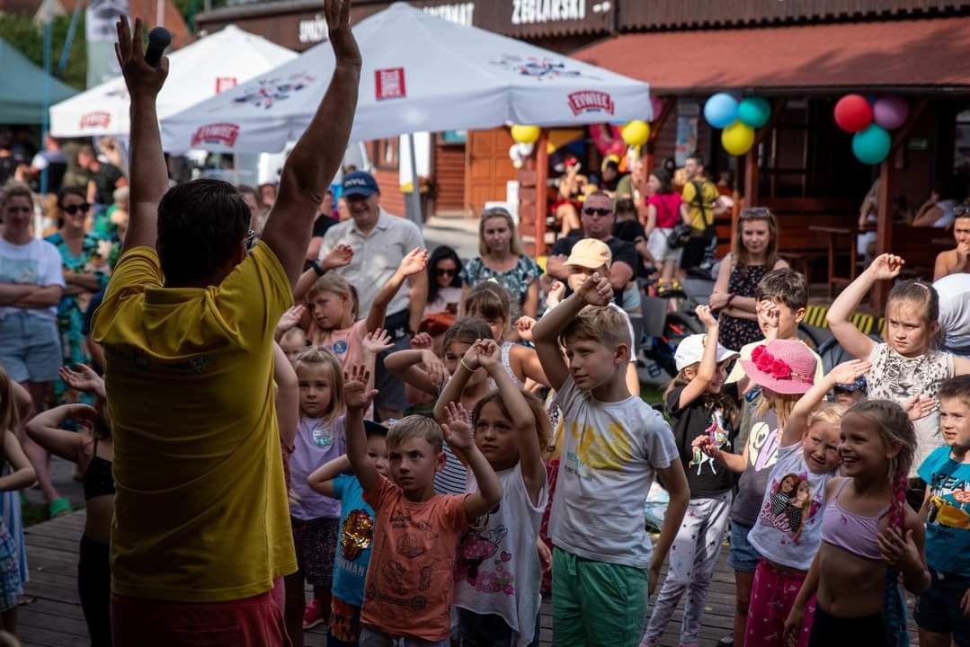 Animator w żółtej koszulce prowadzi grupę dzieci podczas zabawy na świeżym powietrzu, w tle widoczny tłum i dekoracje.