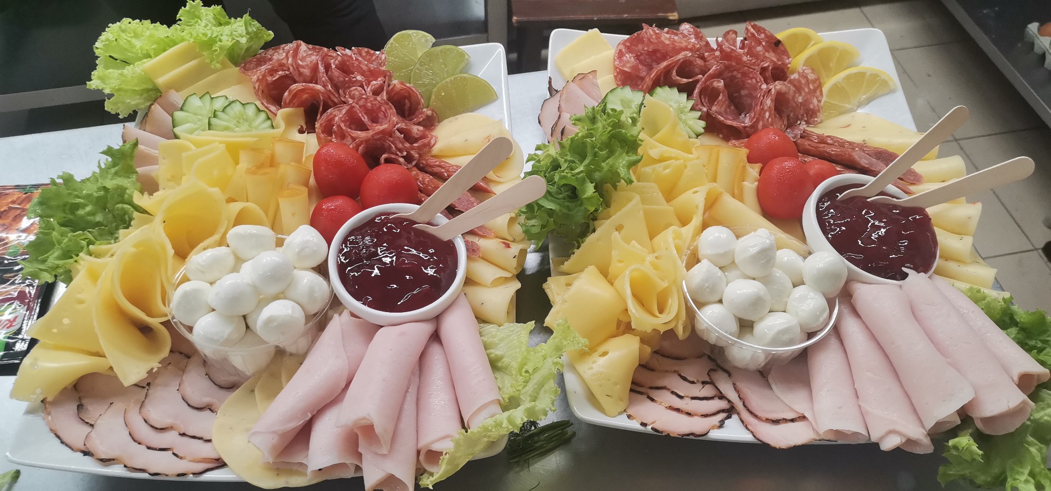 Dwa białe prostokątne talerze z różnorodnymi przekąskami: salami, szynka, kilka rodzajów sera, pomidorki koktajlowe, sałata, ogórki, limonki i miseczka z dżemem z dwoma drewnianymi łyżeczkami.
