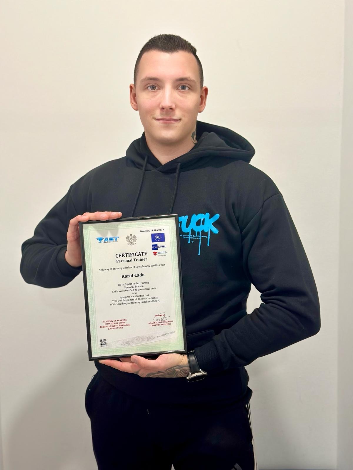 Młody mężczyzna w czarnej bluzie z kapturem prezentuje certyfikat ukończenia kursu trenera personalnego, trzymając go oburącz na neutralnym tle.