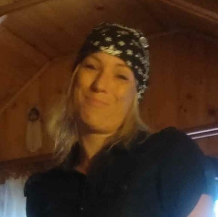 Uśmiechnięta kobieta w czarnej koszuli i bandanie na tle drewnianej ściany w ciepłym oświetleniu.