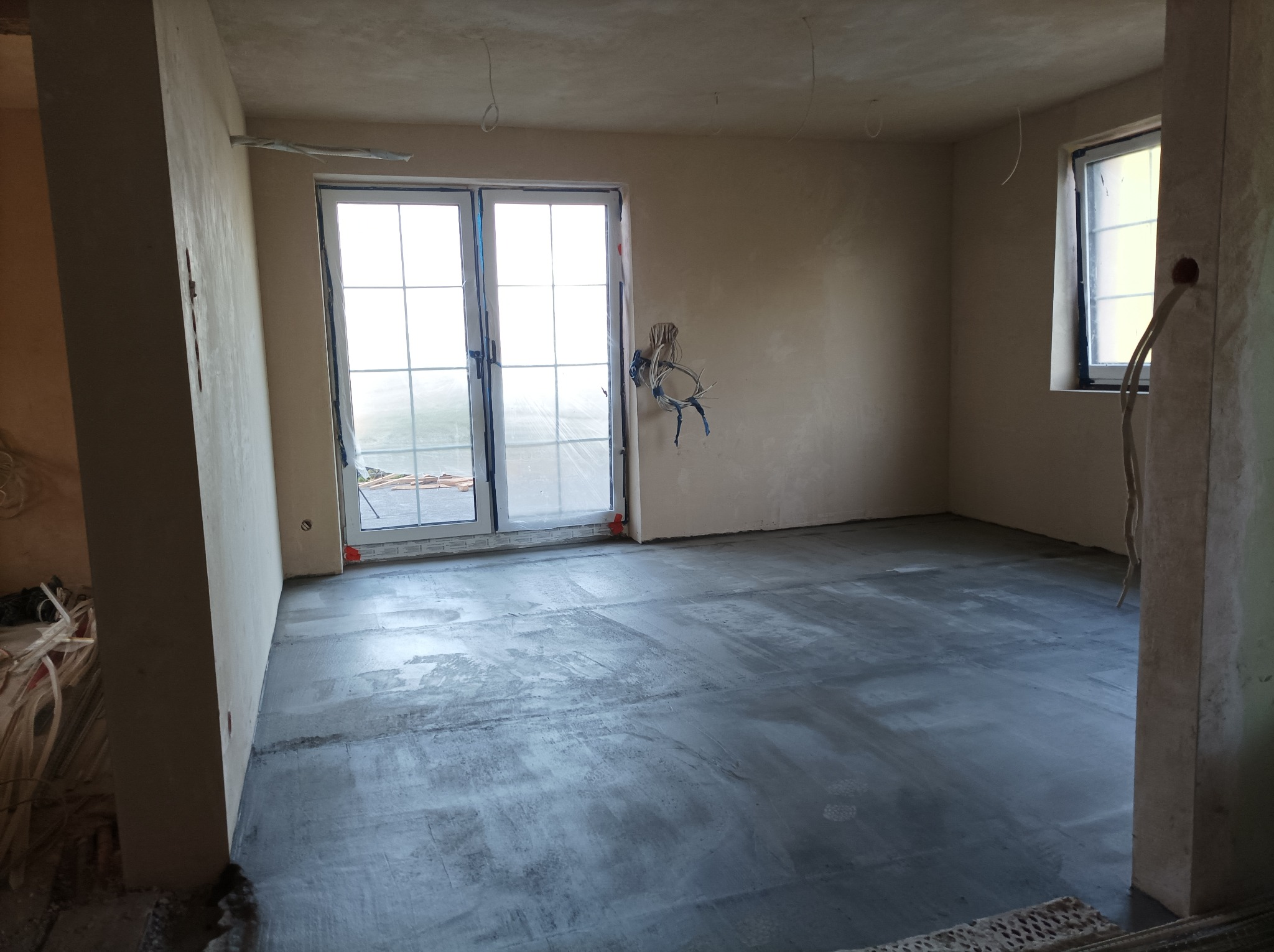 Surowe wnętrze z wylaną, szarą posadzką. Widoczne okno, drzwi balkonowe i wystające kable elektryczne. Ściany przygotowane do dalszych prac wykończeniowych.