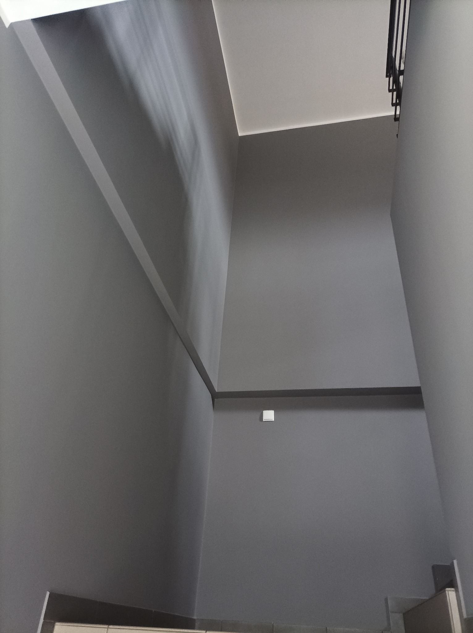 Klatka schodowa po malowaniu na szaro, widok z dołu. Minimalistyczne wykończenie, proste linie, oświetlenie górne. Fragment schodów w dolnej części kadru.