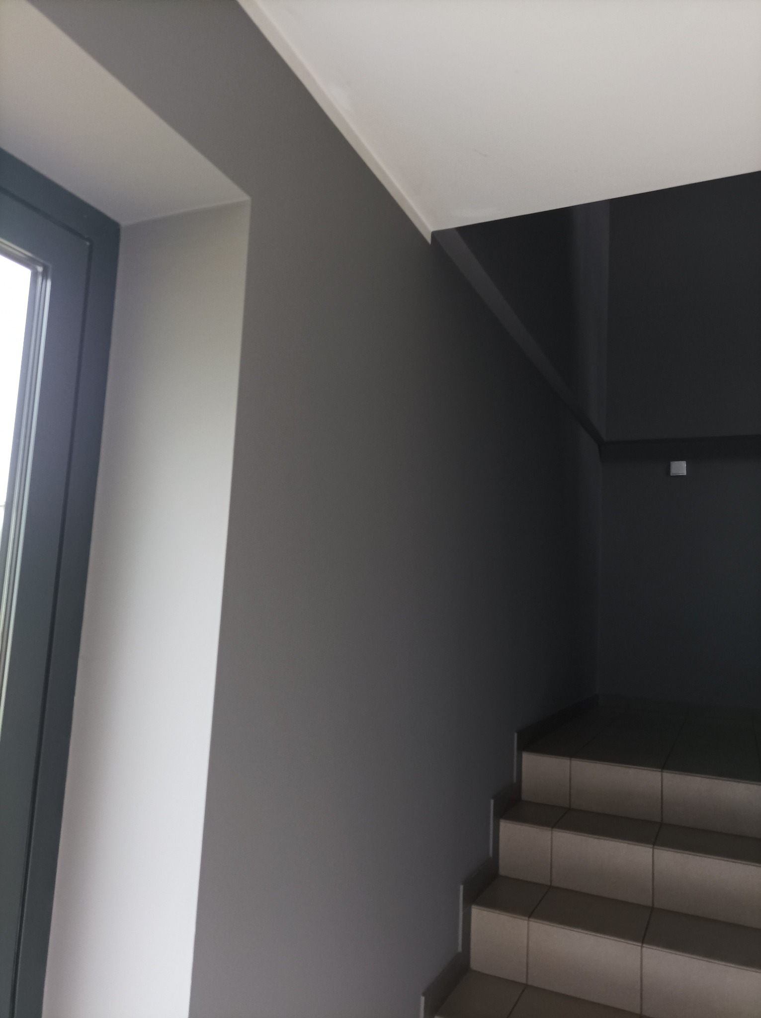 Klatka schodowa w szarościach z oknem po lewej. Minimalistyczny design, płytki na schodach, jednolite kolory ścian. Widok z dołu, neutralne oświetlenie.