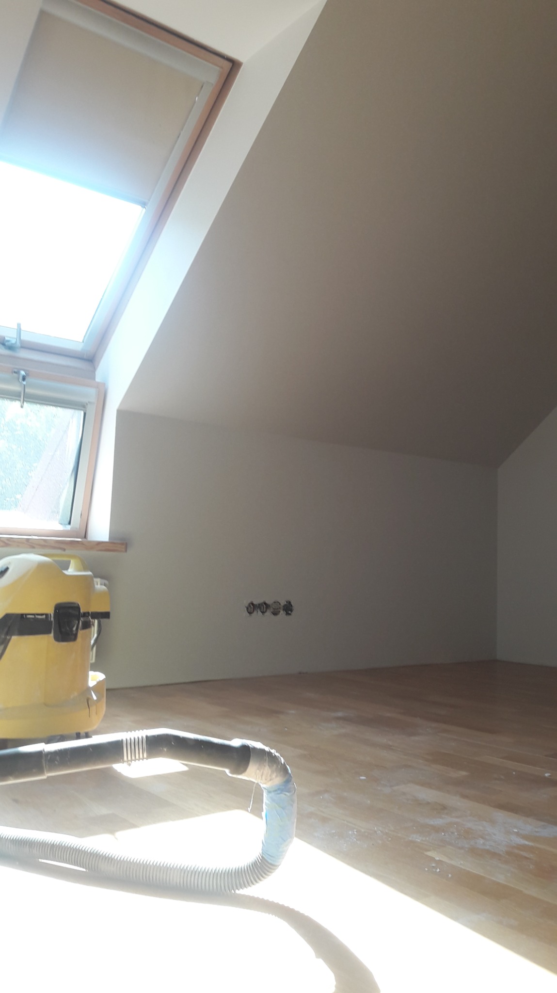 Poddasze po malowaniu ścian na biało, widoczne gniazdka elektryczne i okno dachowe. Na pierwszym planie odkurzacz przemysłowy, co sugeruje sprzątanie po malowaniu.