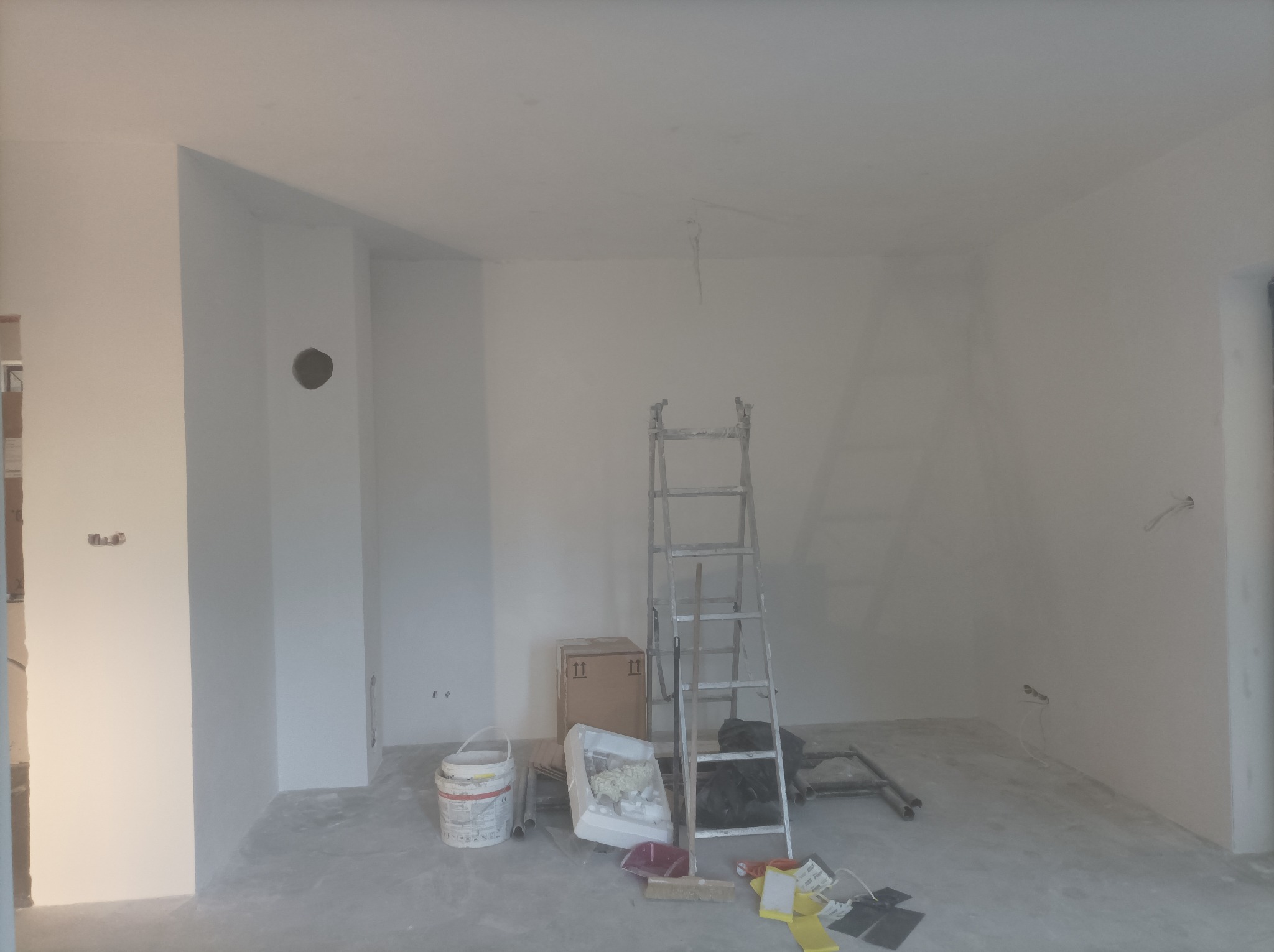 Wnętrze w trakcie malowania: białe ściany, drabina, wiadro z farbą i akcesoria malarskie. Widoczne niedoskonałości i przygotowania do dalszych prac wykończeniowych.