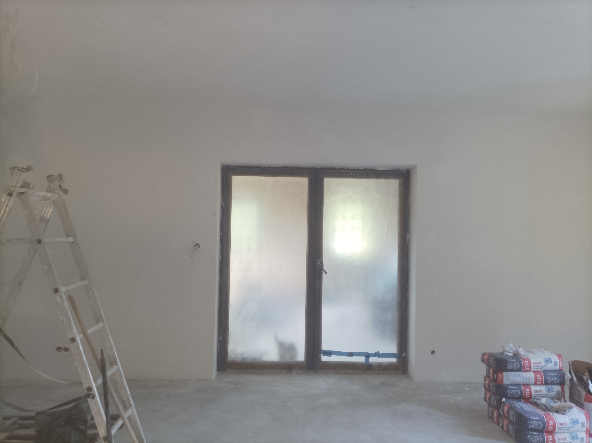 Pokój po gładziowaniu ścian, widoczne worki z gładzią, drabina malarska i okno zabezpieczone folią. Ściany przygotowane do malowania.