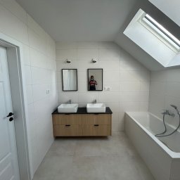 Lordom Sp z o.o. - Jasna łazienka na poddaszu z dwoma umywalkami nablatowymi na szafce z drewnopodobnym frontem, białą wanną i oknem dachowym.
