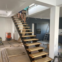 Lordom Sp z o.o. - Montaż drewnianych schodów z czarną konstrukcją w surowym wnętrzu, widoczne zabezpieczenia taśmą malarską, narzędzia i osoby na schodach, kuchnia w tle w trakcie prac wykończeniowych.