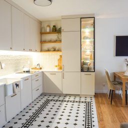 Lordom Sp z o.o. - Jasna kuchnia z białymi szafkami, złotymi uchwytami, wzorzystą podłogą i przeszkloną witryną z oświetleniem LED, połączona z jadalnią z drewnianym stołem i krzesłami, widok na salon.