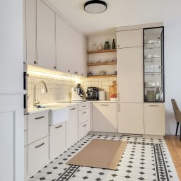 Lordom Sp z o.o. - Jasna kuchnia z białymi frontami szafek, złotymi uchwytami, płytkami w czarno-białe wzory na podłodze i drewnianymi półkami z dekoracjami.