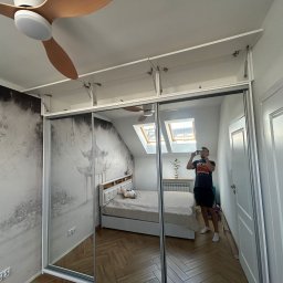 Lordom Sp z o.o. - Szafa wnękowa z lustrzanymi drzwiami przesuwnymi, z nadstawką, odbijająca wnętrze sypialni z łóżkiem i oknem dachowym. Widoczny fragment sufitu z wentylatorem.