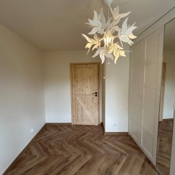 Lordom Sp z o.o. - Jasne wnętrze z parkietem w jodełkę, drewnianymi drzwiami i białą szafą wnękową z lustrem. Dekoracyjna lampa sufitowa w kształcie liści.