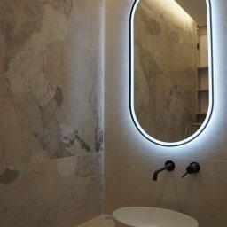 Lordom Sp z o.o. - Elegancka łazienka z owalnym lustrem LED, marmurowymi płytkami i białą umywalką nablatową. Czarna armatura dodaje kontrastu. Widoczne odbicie wnętrza w lustrze.