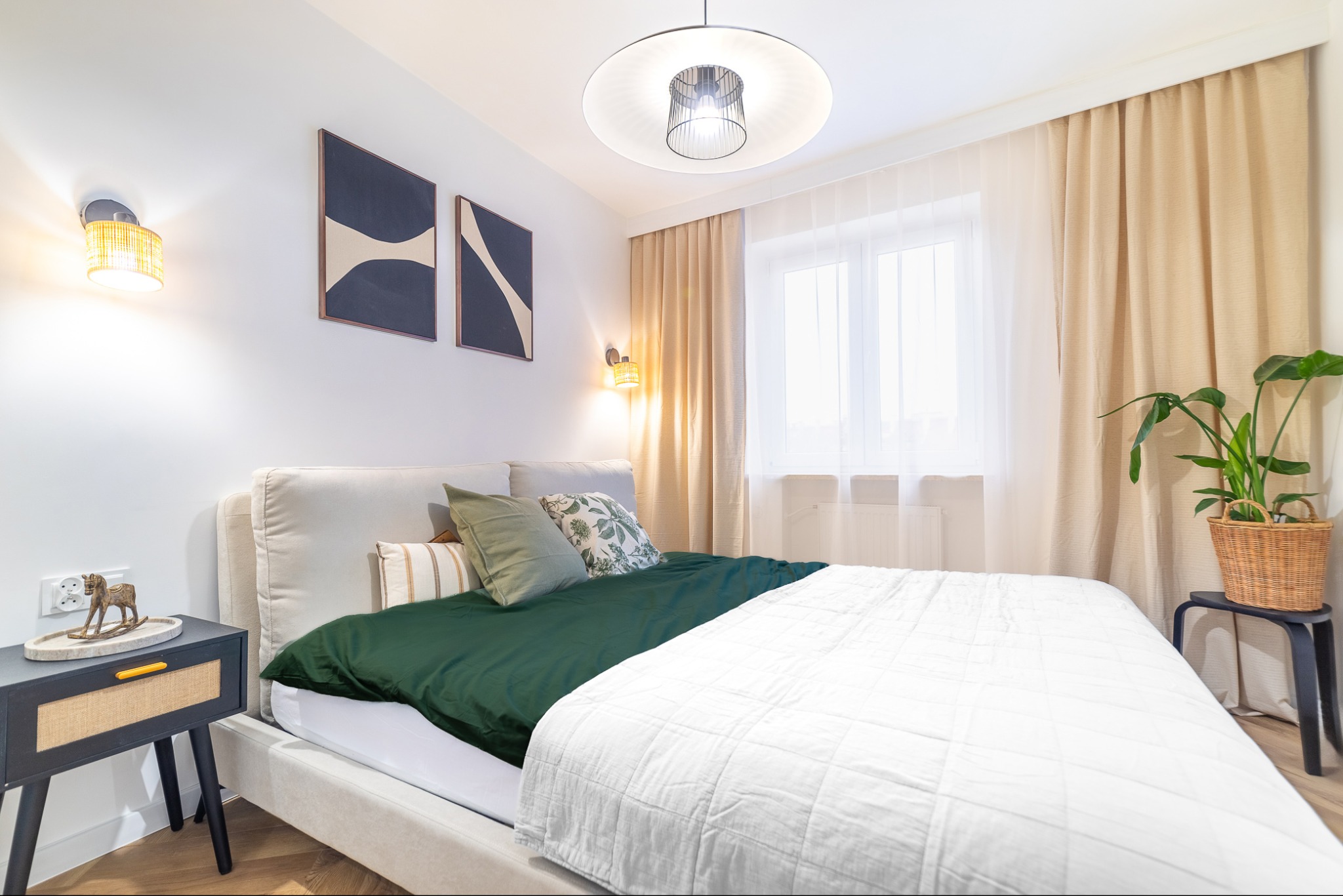 Jasna sypialnia w Krakowie z beżowym łóżkiem, zieloną pościelą, drewnianą szafką nocną z konikiem na blacie, nowoczesnymi lampami ściennymi i wiszącą, oraz zasłonami w oknie.