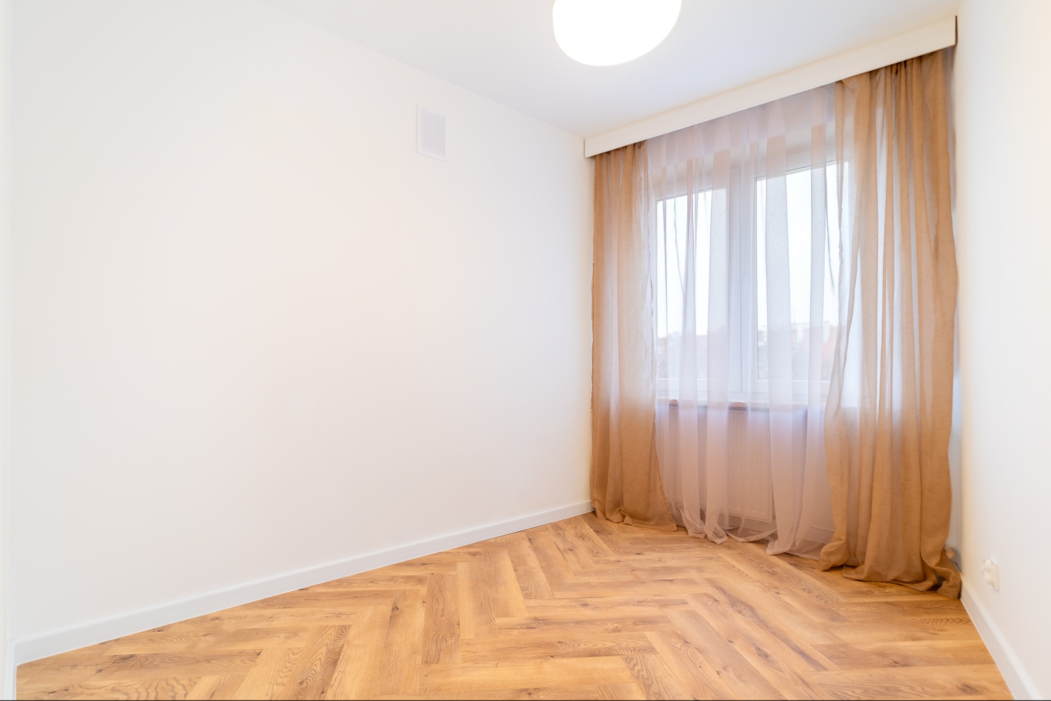 Pusty pokój po remoncie z drewnianą podłogą ułożoną w jodełkę, białymi ścianami, oknem z beżowymi, prześwitującymi zasłonami i okrągłą lampą sufitową.