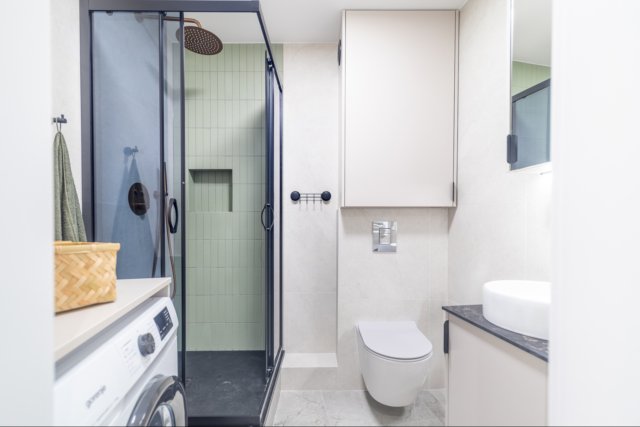 Nowoczesna łazienka z kabiną prysznicową typu walk-in z czarnymi ramami, zielonymi płytkami na ścianie, wiszącą toaletą, umywalką nablatową i pralką pod blatem.