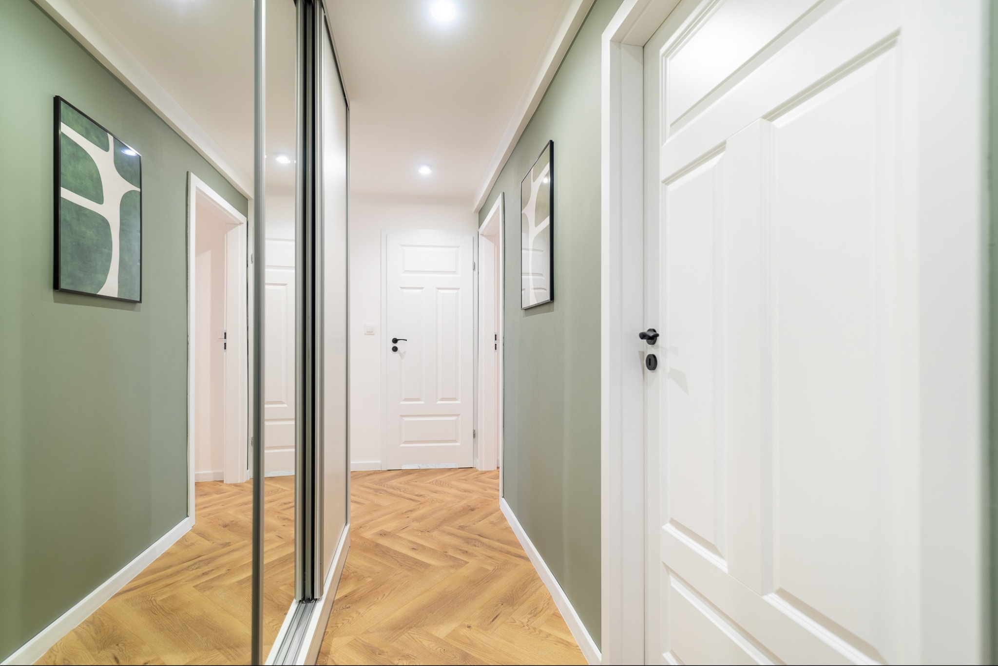 Korytarz w odcieniach zieleni i bieli z podłogą ułożoną w jodełkę, lustrzana szafa przesuwna po lewej stronie, białe drzwi z czarnymi klamkami po prawej, grafika na ścianie.