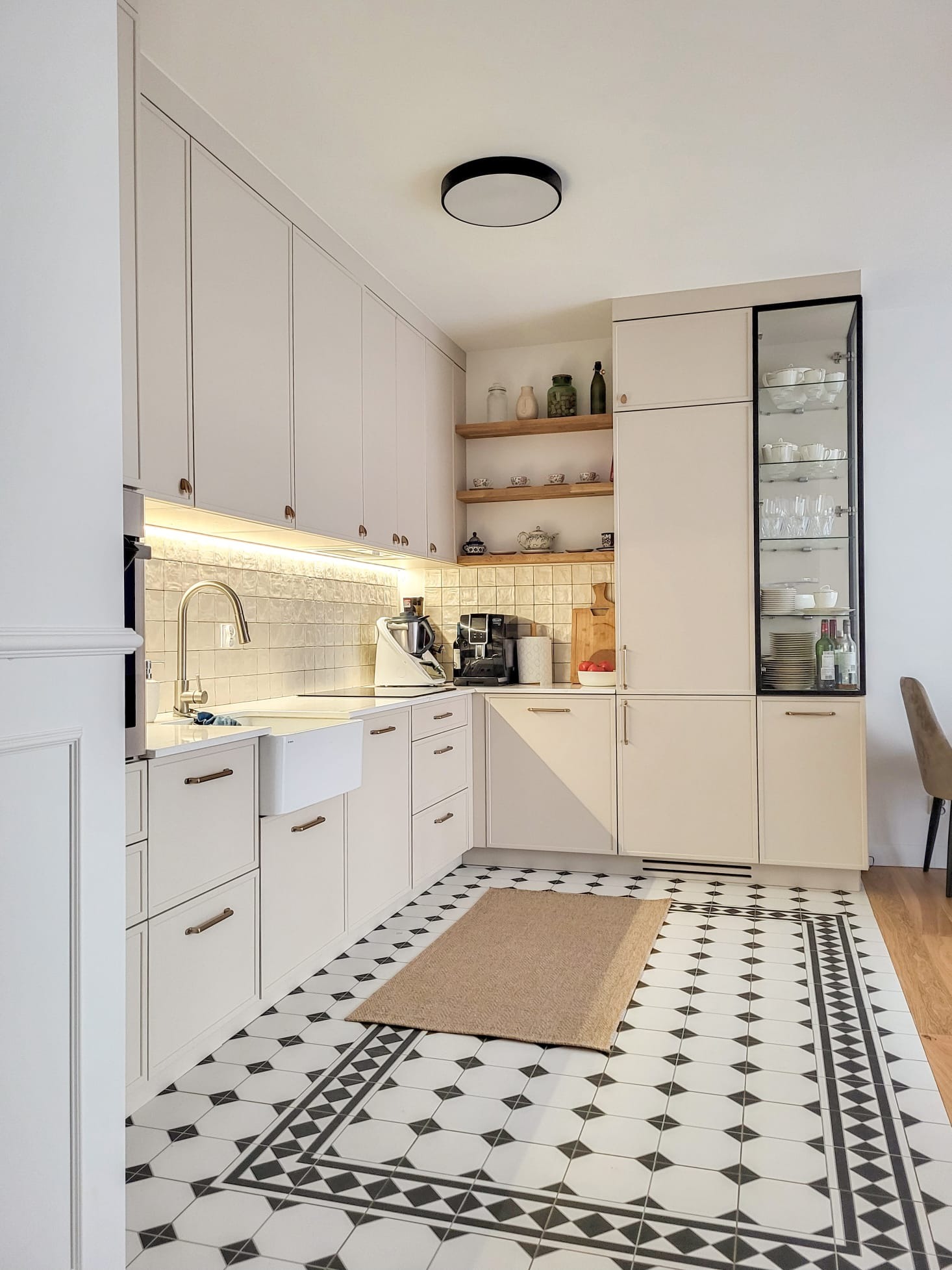Jasna kuchnia z białymi frontami szafek, złotymi uchwytami, płytkami w czarno-białe wzory na podłodze i drewnianymi półkami z dekoracjami.