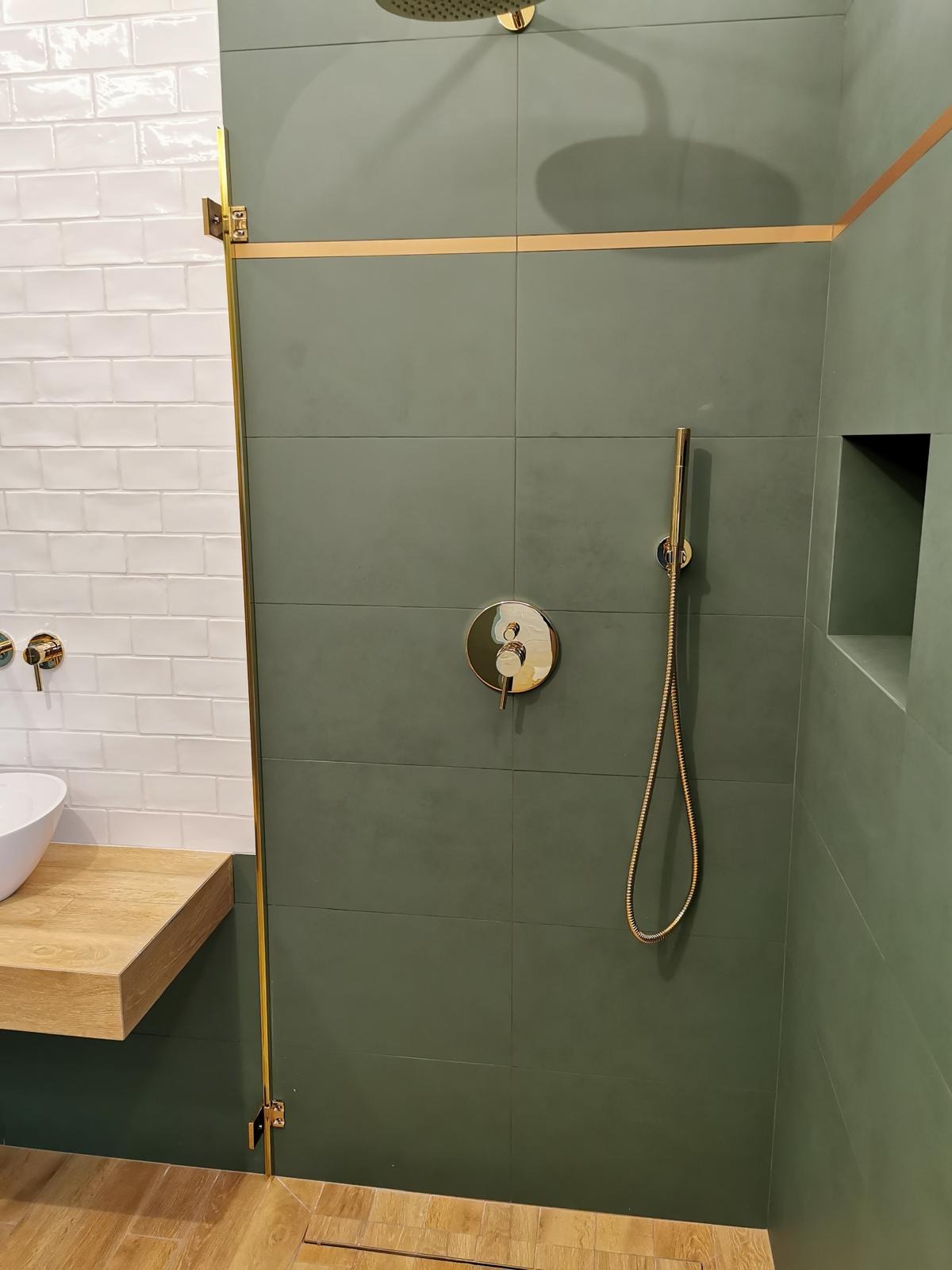 Nowoczesna łazienka z zielonymi płytkami, złotymi akcentami armatury, drewnianym blatem pod umywalką i kabiną prysznicową typu walk-in.