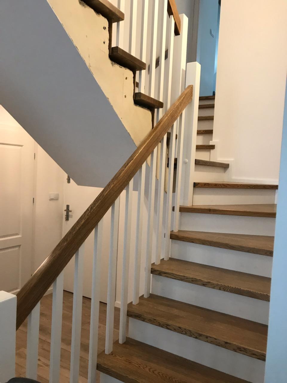 Drewniane schody z białymi balustradami, częściowo w trakcie remontu, ukazujące surowy stan pod stopniami.