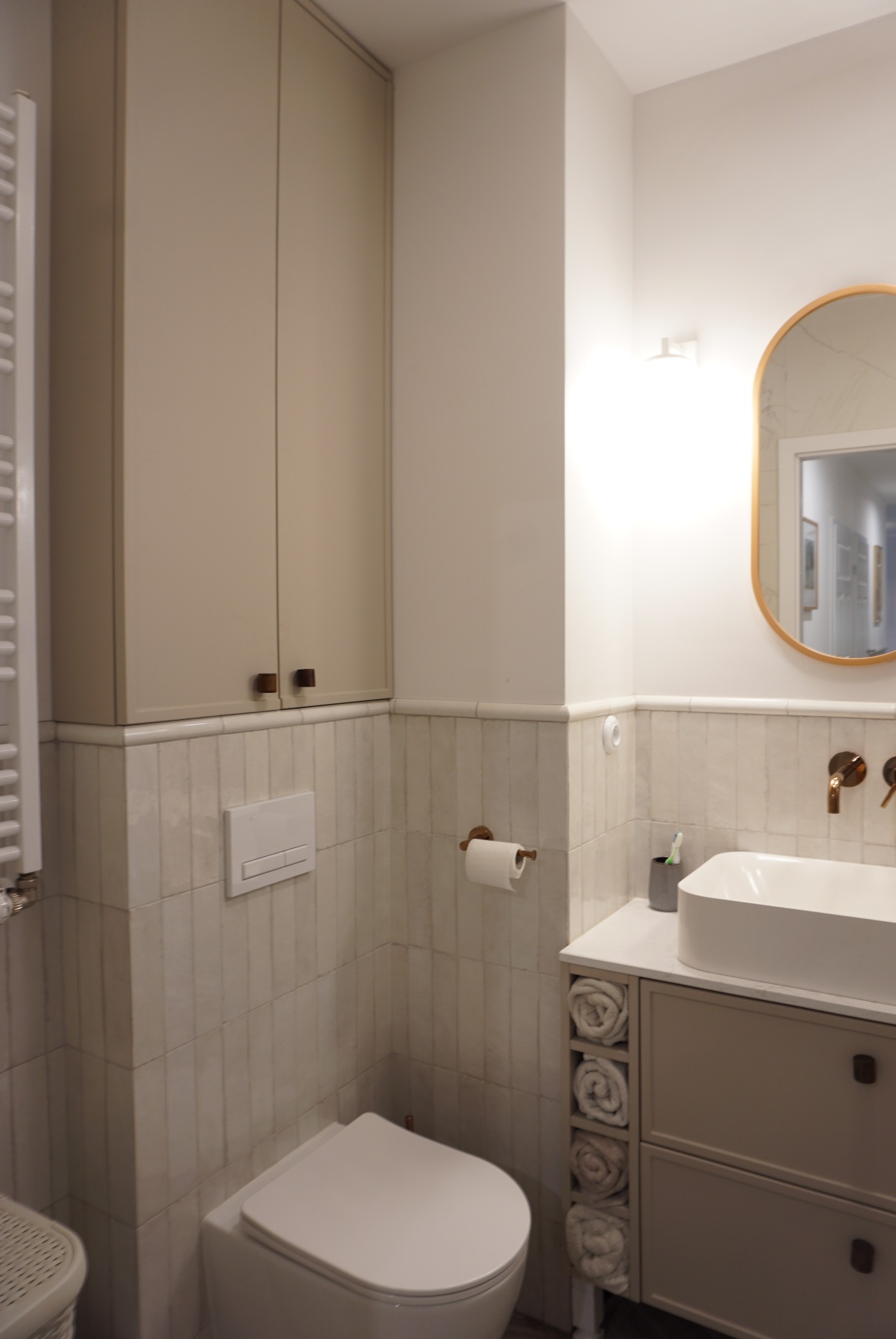 Jasna łazienka z beżowymi szafkami, owalnym lustrem w złotej ramie i toaletą. Ściany wyłożone płytkami imitującymi marmur. Umywalka nablatowa.