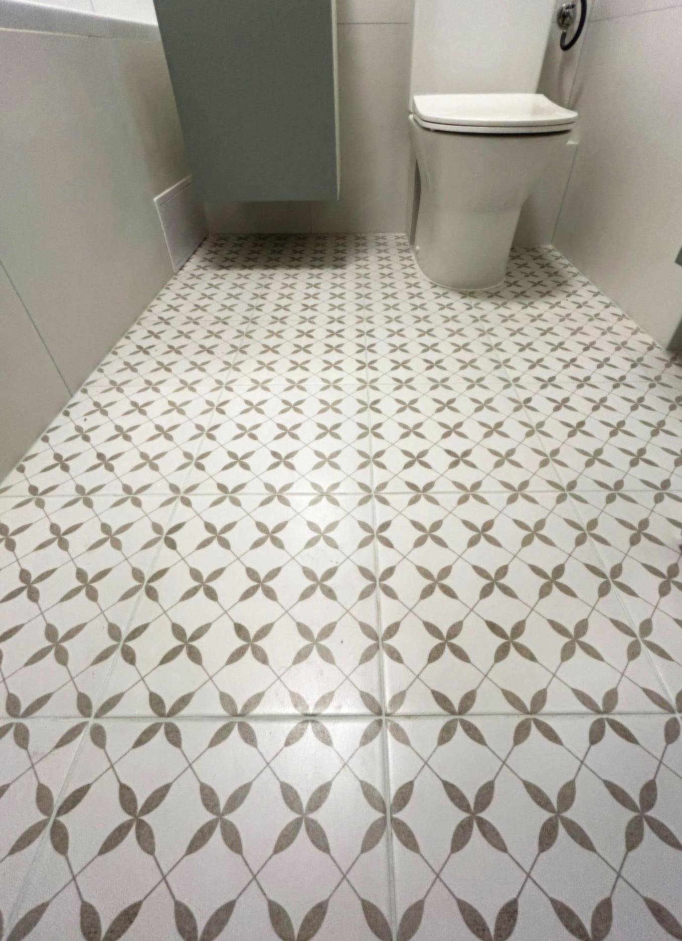 Wzorzysta podłoga w łazience z biało-szarymi płytkami ułożonymi w romby, widoczna toaleta i fragment szafki pod umywalkę. Ujęcie z niskiej perspektywy.