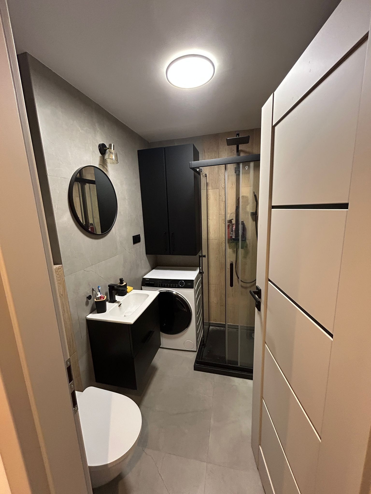 Nowoczesna łazienka z kabiną prysznicową, umywalką z szafką w kolorze czarnym, okrągłym lustrem i pralką. Stonowana kolorystyka, szare płytki, minimalistyczny design.