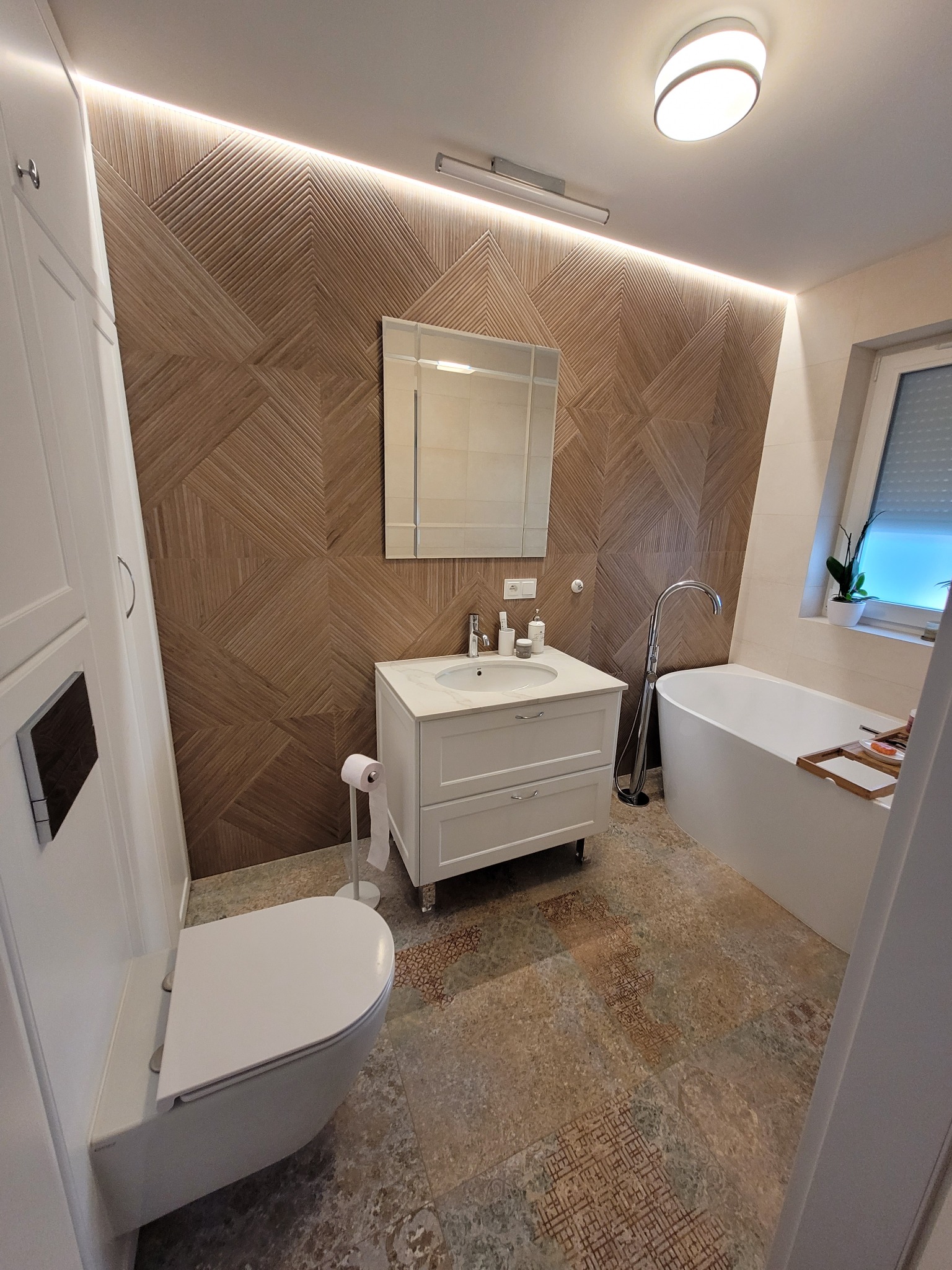 Nowoczesna łazienka z umywalką na postumencie, wolnostojącą wanną i geometryczną okładziną ścienną w odcieniach brązu, oświetlona listwą LED.