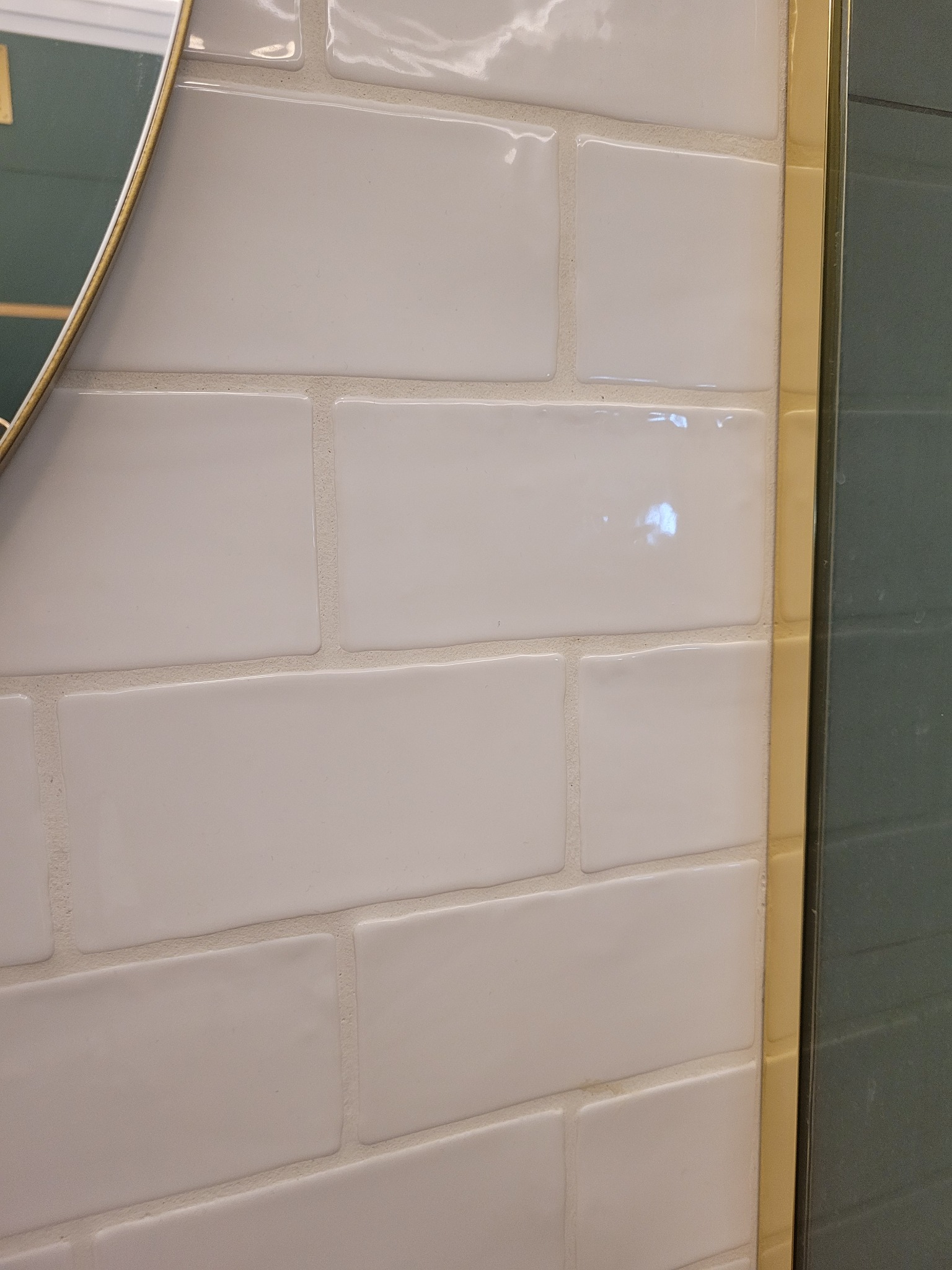 Białe, prostokątne płytki ułożone w cegiełkę, z widoczną złotą ramą lustra i krawędzią kabiny prysznicowej, ukazujące detale wykończenia wnętrza łazienki.